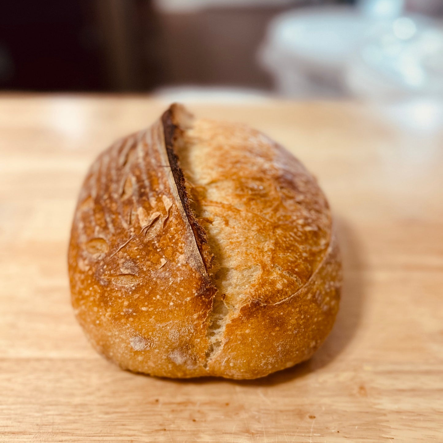 A single rustic round bread roll with a deep scored crack, sitting on a light wooden surface with a softly blurred background.